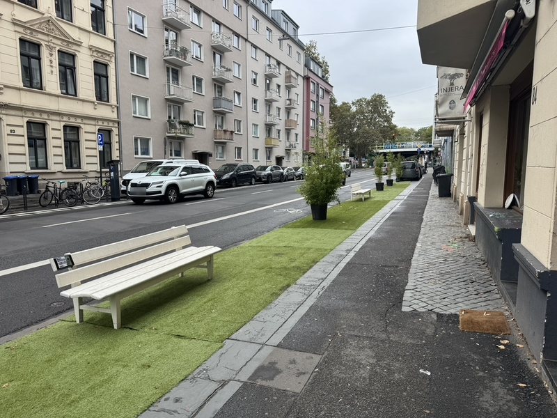Lindenstraße stadtauswärts: Auf dem Gehweg liegt an der Fahrbahnseite ein langer Streifen Kunstrasen. Darauf stehen zwei weiße Sitzbänke und mehrere Pflanzkübel mit Bambus. Daneben verläuft der Radfahrstreifen, der Gehbereich an den Häusern ist frei. Kein Auto steht auf dem Gehweg.